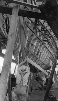 black and white image of man in suspenders and brimmed hat working on the skeleton of Trumpy yacht with framework of boat propped up on supports being helped by another man in the background