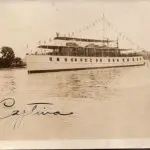 sepia artistic image of trumpy yacht decorated with flags on water with trees on the right side and solid sky in the background, signature in blueish ink on bottom left corner