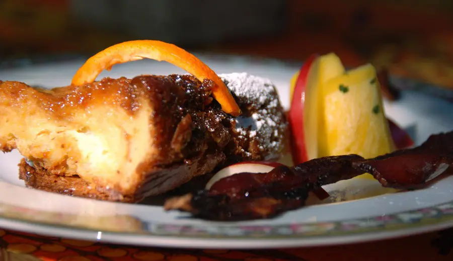 close up of homemade crème Brulee sprinkled with powdered sugar, with pineapple, side of bacon and homemade orange peel with blurred background on white and pink china plate