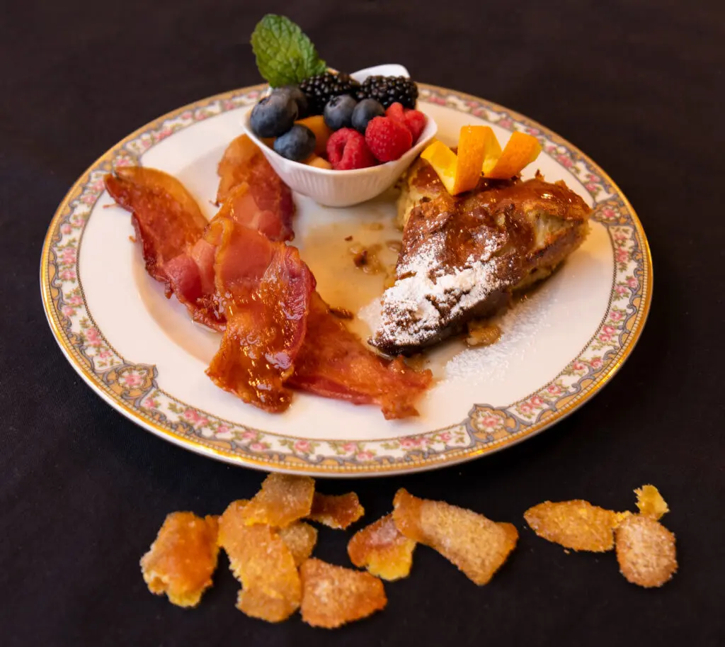 homemade crème Brulee sprinkled with powdered sugar, with mixed fruit cup, side of bacon and homemade orange peel on black background with decorative orange peel in front of china plate