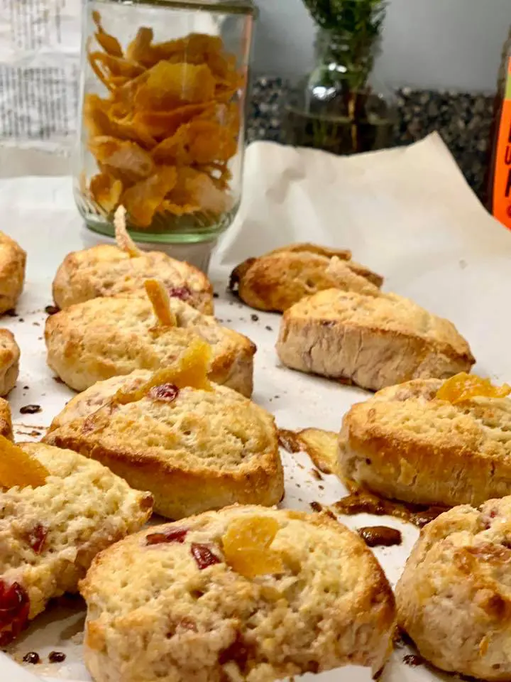 12 homemade orange and craisin scones on baking sheet with jar of homemade orange peels in the back as well as fresh rosemary in a vase and part of a recipe book