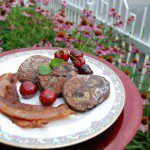 three homemade sourdough banana pancakes with a side of bacon decorated with cherries and powdered sugar on white and pink china plate with red charger against pink flowers with green stems and white picket fence