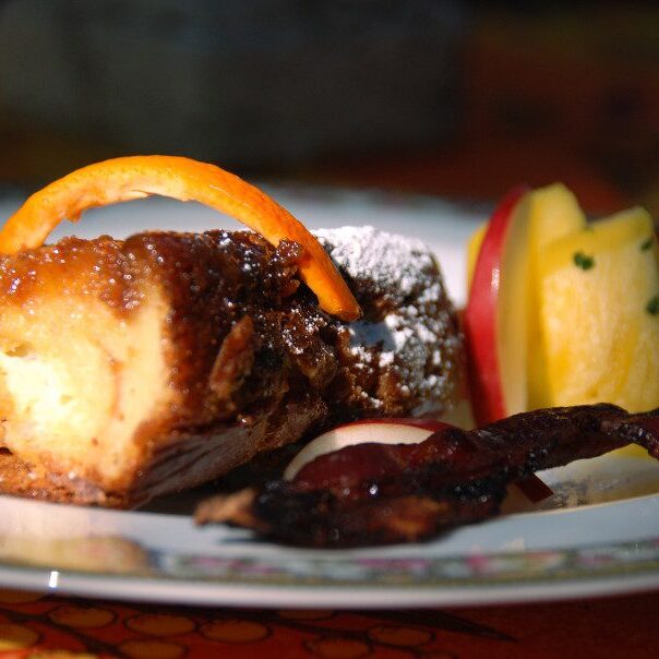 close up of homemade crème Brulee sprinkled with powdered sugar, with pineapple, side of bacon and homemade orange peel with blurred background on white and pink china plate